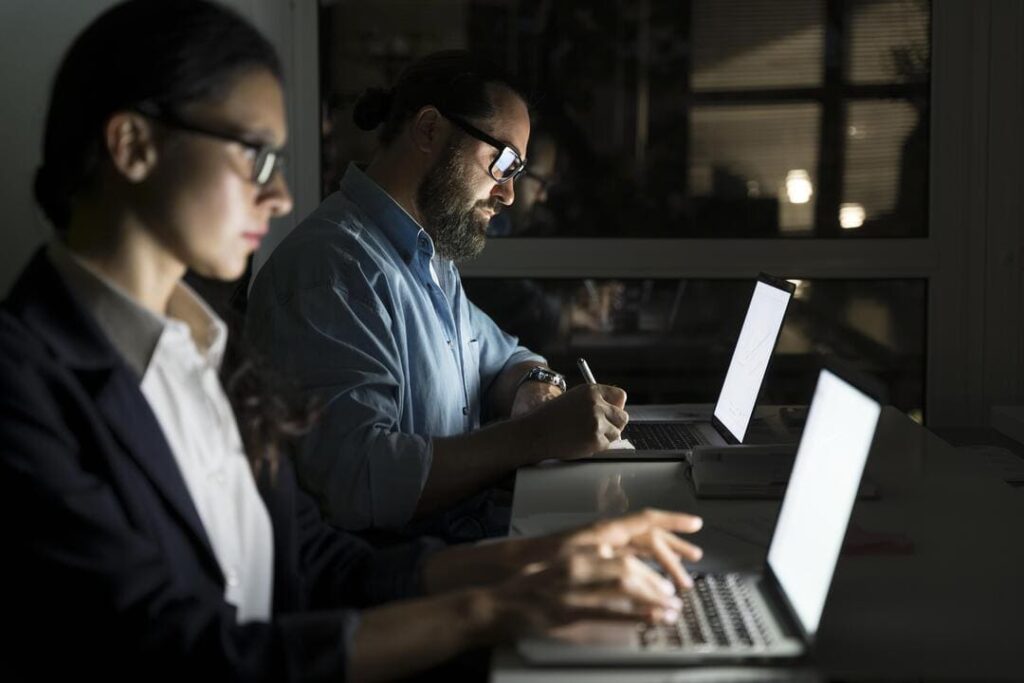 Duas pessoas trabalhando em notebooks à noite, em um ambiente de escritório iluminado apenas pela luz das telas.