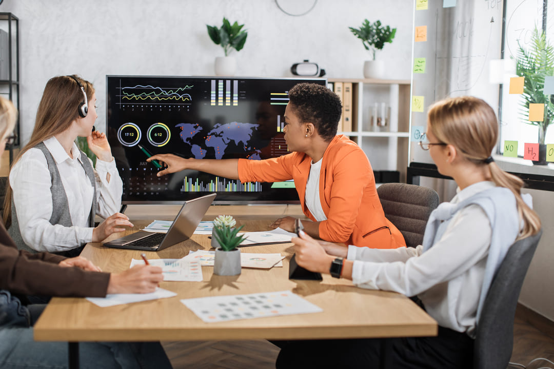Equipe em reunião analisando gráficos e dados em tela grande em ambiente de escritório moderno.