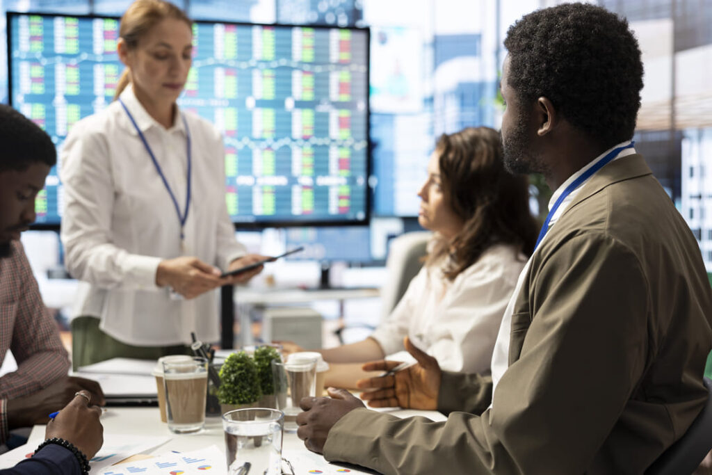 Equipe de profissionais discutindo gráficos e dados em reunião estratégica, com telas ao fundo exibindo indicadores de performance.