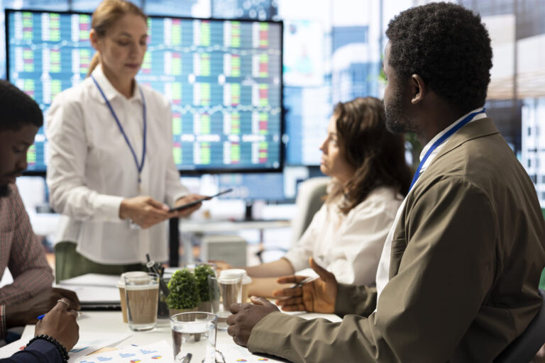 Equipe de profissionais discutindo gráficos e dados em reunião estratégica, com telas ao fundo exibindo indicadores de performance.
